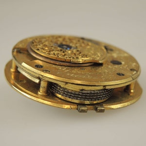 Puede incluir: Un movimiento de reloj de bolsillo dorado con detalles intrincados y una peque&ntilde;a joya azul. El reloj est&aacute; abierto y el mecanismo interno es visible.