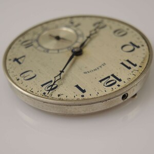 Op de afbeelding: Een zilveren zakhorloge met zwarte wijzers en Romeinse cijfers. Het horloge heeft een licht verweerd gezicht en een klein sub-horloge. Het horloge is gemarkeerd met "Blomquist" op het gezicht.