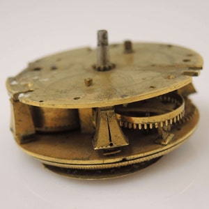 May include: Close-up of a brass clockwork mechanism with intricate gears and a central spindle. The mechanism is disassembled and shows the inner workings of the clock.