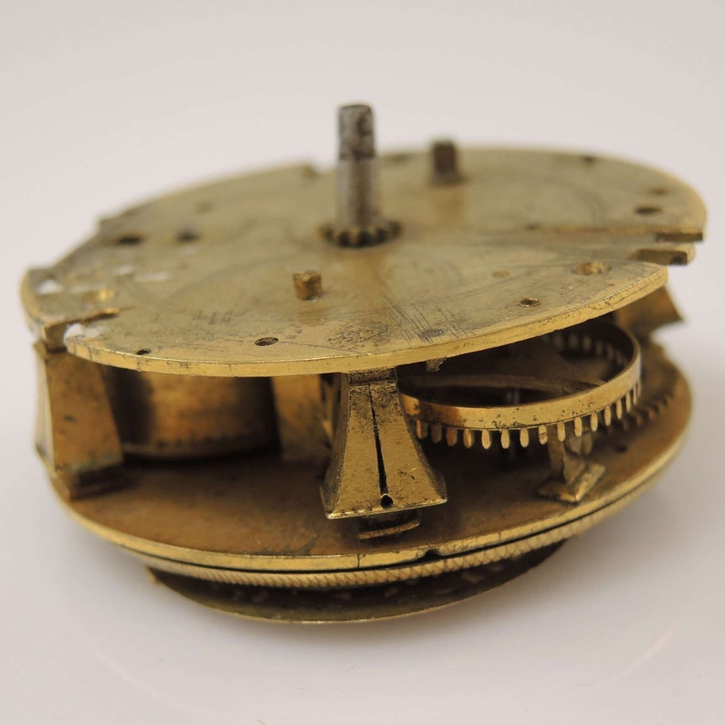 May include: Close-up of a brass clockwork mechanism with intricate gears and a central spindle. The mechanism is disassembled and shows the inner workings of the clock.