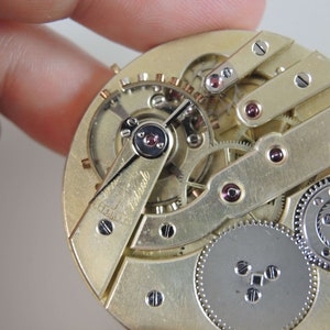 Puede incluir: Primer plano de un movimiento de reloj de bolsillo de tono dorado con engranajes intrincados y un engranaje de tono plateado en la esquina inferior derecha. El movimiento est&aacute; etiquetado como "Remontoir".