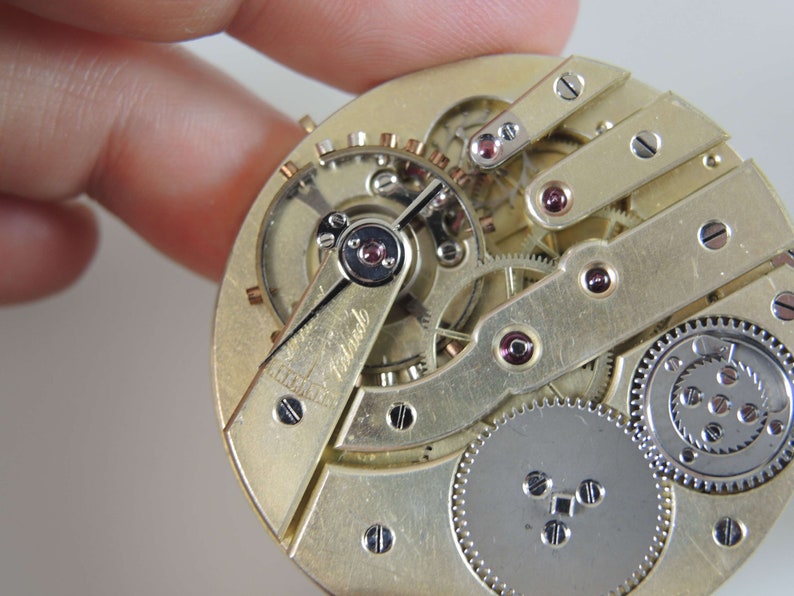 Puede incluir: Primer plano de un movimiento de reloj de bolsillo de tono dorado con engranajes intrincados y un engranaje de tono plateado en la esquina inferior derecha. El movimiento est&aacute; etiquetado como "Remontoir".