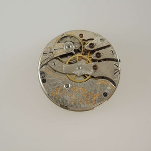 Op de afbeelding: Close-up van de binnenkant van een zilveren zakhorloge. De wijzerplaat van het horloge is versierd met een ingewikkeld ontwerp en het mechanisme is zichtbaar.