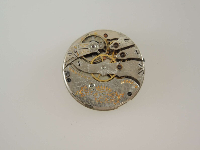 Op de afbeelding: Close-up van de binnenkant van een zilveren zakhorloge. De wijzerplaat van het horloge is versierd met een ingewikkeld ontwerp en het mechanisme is zichtbaar.