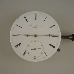 Op de afbeelding: Een wit zakhorloge-wijzerplaat met zwarte Romeinse cijfers voor de uren en zwarte markeringen voor de minuten. De wijzerplaat van het horloge heeft een kleinere witte sub-wijzerplaat met zwarte markeringen voor de minuten. Op de wijzerplaat van het horloge staat de tekst "Ulisse Nardin, Le Locle". De wijzerplaat van het horloge is bevestigd aan een goudkleurige opwindsleutel.