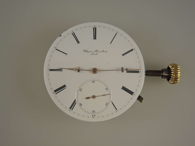 Op de afbeelding: Een wit zakhorloge-wijzerplaat met zwarte Romeinse cijfers voor de uren en zwarte markeringen voor de minuten. De wijzerplaat van het horloge heeft een kleinere witte sub-wijzerplaat met zwarte markeringen voor de minuten. Op de wijzerplaat van het horloge staat de tekst "Ulisse Nardin, Le Locle". De wijzerplaat van het horloge is bevestigd aan een goudkleurige opwindsleutel.