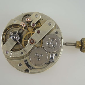 May include: Close-up of a silver watch movement with intricate gears and a winding stem. The movement is exposed, showing the inner workings of the watch.
