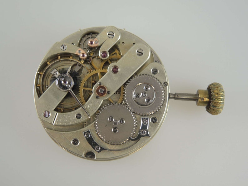 May include: Close-up of a silver watch movement with intricate gears and a winding stem. The movement is exposed, showing the inner workings of the watch.