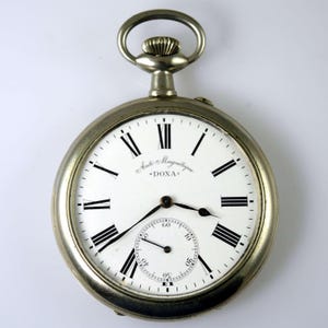 May include: A silver-toned pocket watch with a white dial, black Roman numeral hour markers, and black hands. The face features the text Anti-Magnétique and DOXA. It includes a small sub-dial for seconds at the bottom and a winding crown at the top.