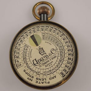 Puede incluir: Una guía de exposición circular vintage para cámara, fabricada por Adams & Co. en Londres. La guía presenta un dial giratorio con diafragmas y velocidades de obturación, junto con una tabla para diferentes sensibilidades de película. La guía está hecha de latón y tiene un diseño en blanco y negro.