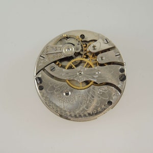 Puede incluir: Primer plano del mecanismo interno de un reloj de bolsillo de plata con engranajes dorados. La esfera del reloj est&aacute; grabada con un dise&ntilde;o floral.