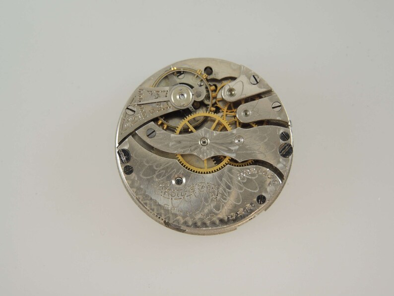 Puede incluir: Primer plano del mecanismo interno de un reloj de bolsillo de plata con engranajes dorados. La esfera del reloj est&aacute; grabada con un dise&ntilde;o floral.