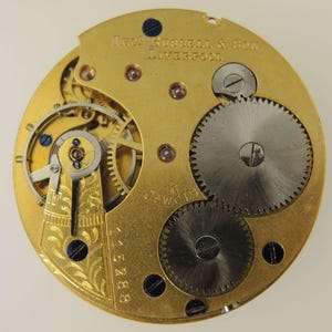 May include: Close-up of a vintage watch movement with intricate gears and components. The gold-toned metal base features the text "Thos. Russell & Son. Liverpool." and "16 Jewels." The watch mechanism includes small screws and blue accents.