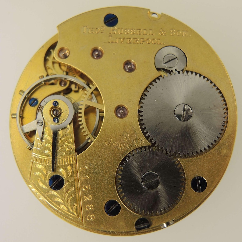 May include: Close-up of a vintage watch movement with intricate gears and components. The gold-toned metal base features the text "Thos. Russell & Son. Liverpool." and "16 Jewels." The watch mechanism includes small screws and blue accents.