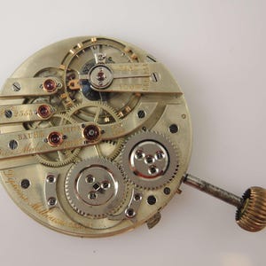 Puede incluir: Un movimiento de reloj de bolsillo de plata con engranajes intrincados y un v&aacute;stago de cuerda. El reloj est&aacute; etiquetado como "Baume & Co. Gen&egrave;ve" y "Prix M&eacute;daille d'Or" con el n&uacute;mero de serie "25632".