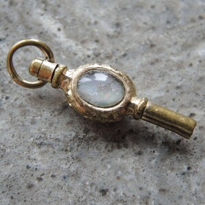 May include: A gold-toned pocket watch key with a round, clear glass face. The glass has a cloudy, iridescent appearance.