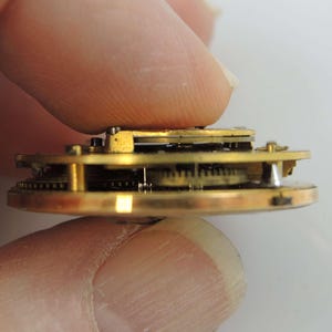 Puede incluir: Vista lateral de cerca de un mecanismo de reloj de bolsillo de tono dorado con engranajes intrincados y una placa de lat&oacute;n. El reloj se sostiene en una mano.