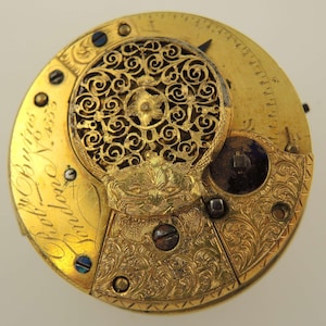 Puede incluir: Primer plano de un movimiento de reloj antiguo con intrincados detalles de filigrana dorada y grabados. El mecanismo del reloj presenta un dise&ntilde;o circular con engranajes, tornillos y un patr&oacute;n floral decorativo visibles. El texto "Robt. Burris London" est&aacute; inscrito.