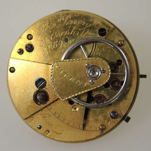Puede incluir: Primer plano del mecanismo interno de un reloj de bolsillo de oro. La esfera del reloj est&aacute; inscrita con "Mr. Lowry & Son, Cornhill, London, 1889".