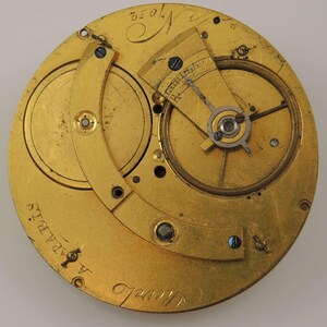 May include: Close-up of a gold-colored pocket watch movement with the inscription "Alfred Chapuis" and "Paris" visible. The watch has a circular design with intricate gears and a lever.