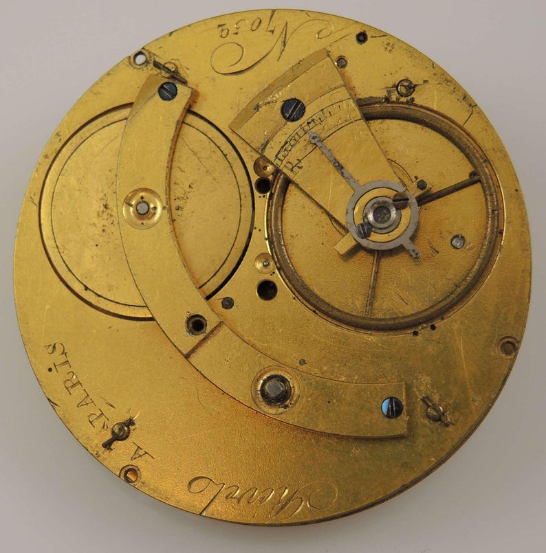 May include: Close-up of a gold-colored pocket watch movement with the inscription "Alfred Chapuis" and "Paris" visible. The watch has a circular design with intricate gears and a lever.