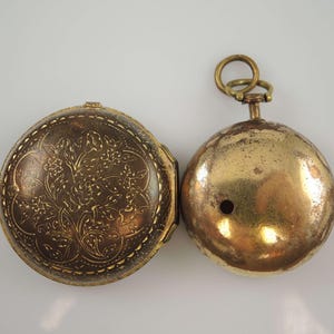 Könnte beinhalten: Eine goldene Taschenuhr mit einem floralen Design auf der Vorderseite und einer schlichten Rückseite. Die Uhr hat einen Klappdeckel und einen Ring zum Anbringen einer Kette.