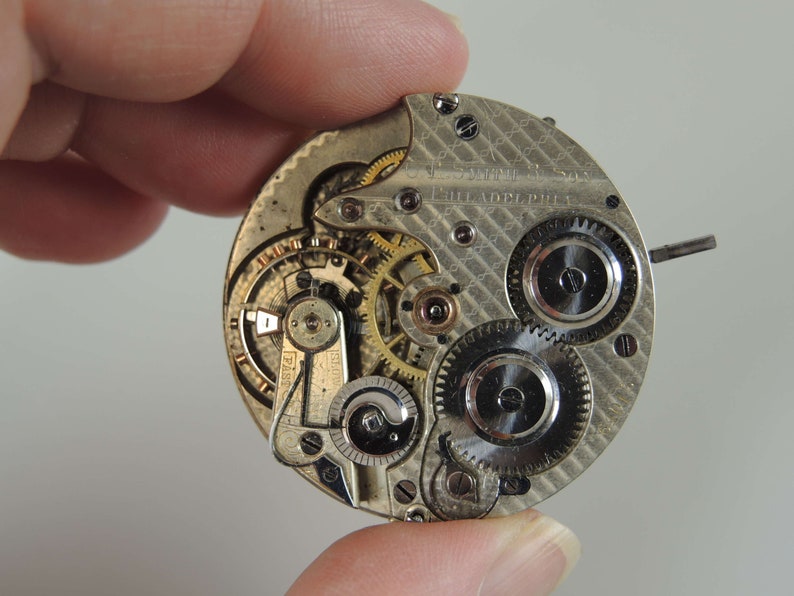 May include: Close-up of a silver pocket watch movement with intricate gears and a patterned back plate. The watch is labeled "J.S. DIME & SON PHILADELPHIA".
