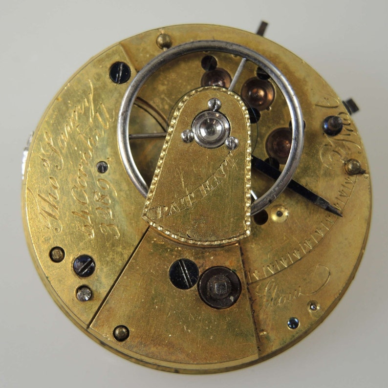 Puede incluir: Primer plano de un movimiento de reloj de bolsillo de oro con la inscripci&oacute;n "The Sentry 14 Carat 1859" y "Patent Henry" visible. El reloj tiene una corona de cuerda plateada y una aguja negra.