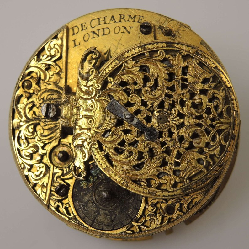 May include: A gold pocket watch with intricate, engraved floral designs. The watch face is visible and features the inscription "De Charme London".