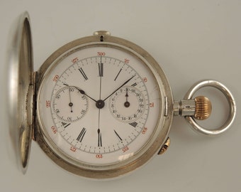 May include: A silver pocket watch with a white face and black hands. The watch has a chronograph function with a smaller dial at the top. The watch is open and the case is visible.