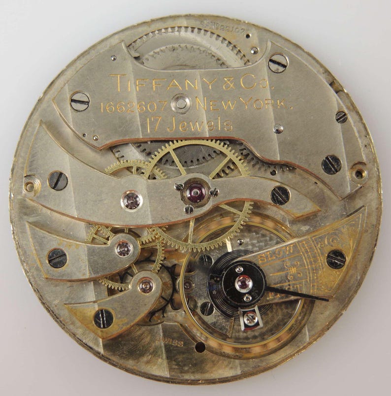 K&ouml;nnte beinhalten: Nahaufnahme eines silbernen Taschenuhrwerks mit goldenen Zahnr&auml;dern und einem schwarzen Aufzugstift. Die Uhr ist mit "Tiffany & Co. 1662607 New York 17 Jewels" beschriftet.