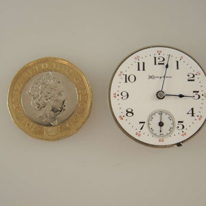 Op de afbeelding: Een wit zakhorloge met zwarte wijzers en een kleine secondewijzer-wijzerplaat. De wijzerplaat van het horloge heeft zwarte Romeinse cijfers als uurmarkeringen en zwarte minutenmarkeringen. Het horloge is omgeven door een zilverkleurige kast.