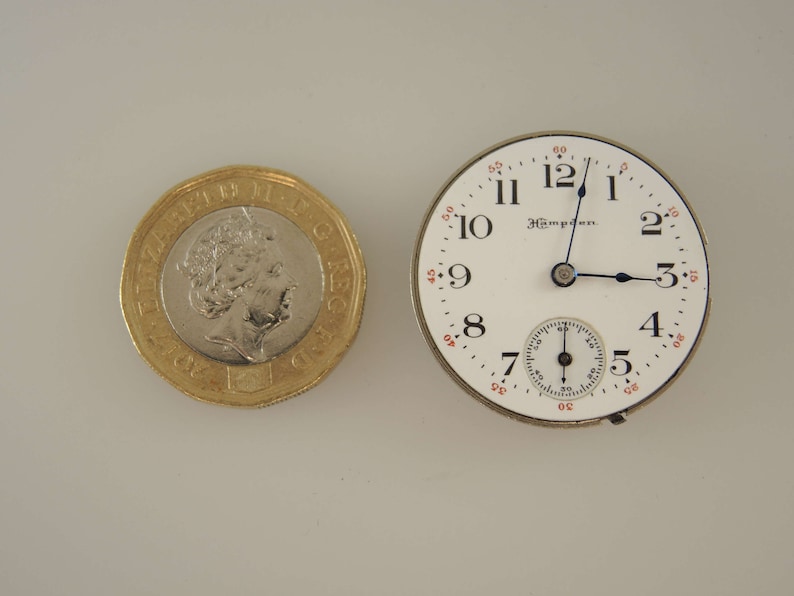Op de afbeelding: Een wit zakhorloge met zwarte wijzers en een kleine secondewijzer-wijzerplaat. De wijzerplaat van het horloge heeft zwarte Romeinse cijfers als uurmarkeringen en zwarte minutenmarkeringen. Het horloge is omgeven door een zilverkleurige kast.