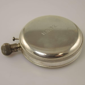 K&ouml;nnte beinhalten: Eine silberfarbene, runde Flasche mit einem strukturierten, geriffelten Verschluss und einem kleinen, zylindrischen Ausguss. Die Flasche hat einen Klappdeckel und ist mit den Initialen "H.S. T2" graviert. Die Flasche ist wahrscheinlich aus Metall.