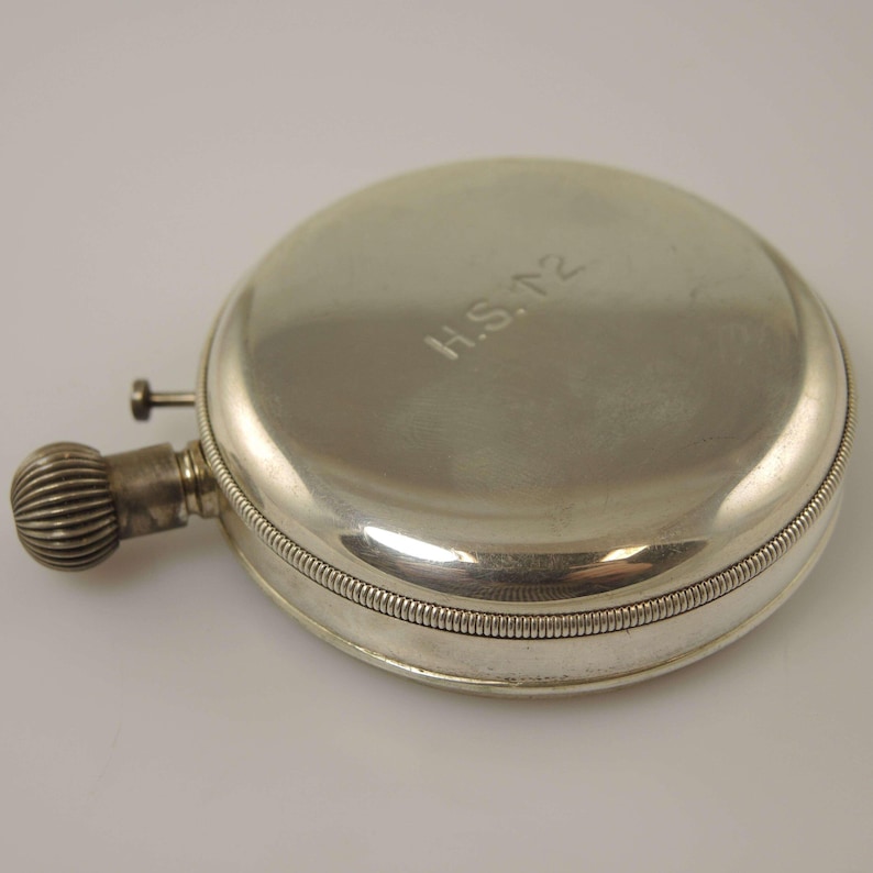 K&ouml;nnte beinhalten: Eine silberfarbene, runde Flasche mit einem strukturierten, geriffelten Verschluss und einem kleinen, zylindrischen Ausguss. Die Flasche hat einen Klappdeckel und ist mit den Initialen "H.S. T2" graviert. Die Flasche ist wahrscheinlich aus Metall.