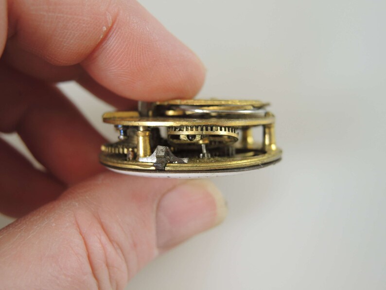 K&ouml;nnte beinhalten: Nahaufnahme eines goldfarbenen Taschenuhrwerks mit komplizierten Zahnr&auml;dern und einem wei&szlig;en Emaille-Zifferblatt.