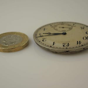 K&ouml;nnte beinhalten: Eine silberne Taschenuhr mit schwarzen r&ouml;mischen Ziffern auf dem Zifferblatt. Die Uhr ist etwas kleiner als eine britische Pfundm&uuml;nze.