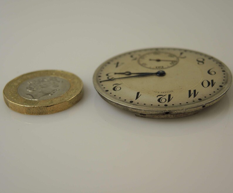 K&ouml;nnte beinhalten: Eine silberne Taschenuhr mit schwarzen r&ouml;mischen Ziffern auf dem Zifferblatt. Die Uhr ist etwas kleiner als eine britische Pfundm&uuml;nze.