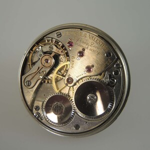 K&ouml;nnte beinhalten: Nahaufnahme eines silbernen Taschenuhrwerks mit komplizierten Zahnr&auml;dern und einer goldenen Inschrift mit dem Text "Lange & Valentine, Swiss Levers, Fabrique Eleyta, 6 rubis".