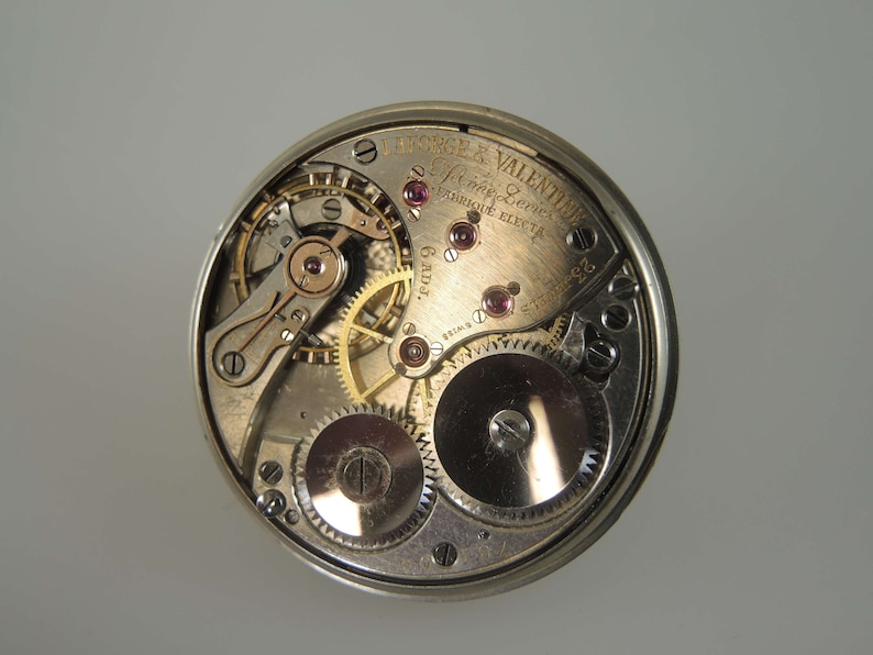 K&ouml;nnte beinhalten: Nahaufnahme eines silbernen Taschenuhrwerks mit komplizierten Zahnr&auml;dern und einer goldenen Inschrift mit dem Text "Lange & Valentine, Swiss Levers, Fabrique Eleyta, 6 rubis".