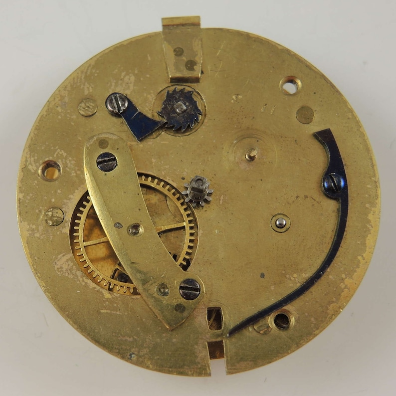 May include: A close-up view of the back of a gold-colored pocket watch movement. The movement has several gears and levers, including a blue lever and a gold-colored lever. The movement is set against a white background.