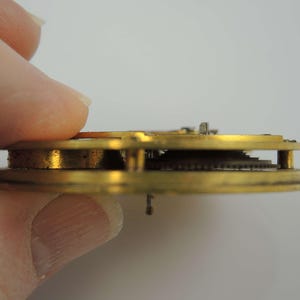 May include: Close-up side view of a gold-colored watch movement with visible gears and a brass frame.