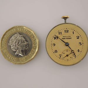 K&ouml;nnte beinhalten: Eine Vintage-Taschenuhr mit cremefarbenem Zifferblatt, schwarzen Ziffern und Zeigern. Auf dem Zifferblatt steht "HAMILTON MARY BIGHAM". Neben der Uhr liegt eine britische Ein-Pfund-M&uuml;nze. Die Uhr hat ein kleines Sekunden-Zifferblatt.