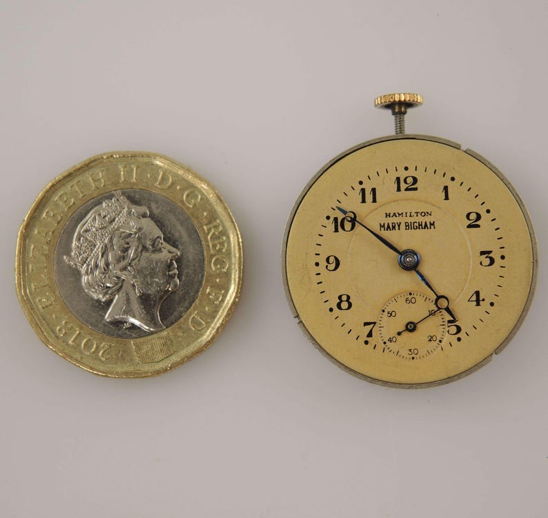 K&ouml;nnte beinhalten: Eine Vintage-Taschenuhr mit cremefarbenem Zifferblatt, schwarzen Ziffern und Zeigern. Auf dem Zifferblatt steht "HAMILTON MARY BIGHAM". Neben der Uhr liegt eine britische Ein-Pfund-M&uuml;nze. Die Uhr hat ein kleines Sekunden-Zifferblatt.