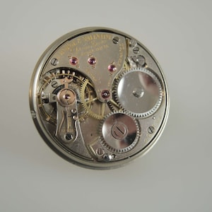K&ouml;nnte beinhalten: Nahaufnahme des Innenlebens einer silbernen Taschenuhr. Das Zifferblatt der Uhr ist mit dem Text "Waltham  Valentine  17 Jewels  10 Size  Adjusted  to Temperature  and  Five Positions" sichtbar.