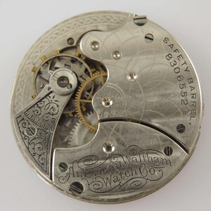 May include: Close-up of a vintage watch movement. The silver-toned mechanism features intricate gears, screws, and engraved details. The text "SAFETY BARREL 8306552" and "American Waltham Watch Co." are visible. The watch movement is a detailed example of early watchmaking.