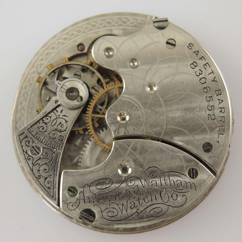 May include: Close-up of a vintage watch movement. The silver-toned mechanism features intricate gears, screws, and engraved details. The text "SAFETY BARREL 8306552" and "American Waltham Watch Co." are visible. The watch movement is a detailed example of early watchmaking.
