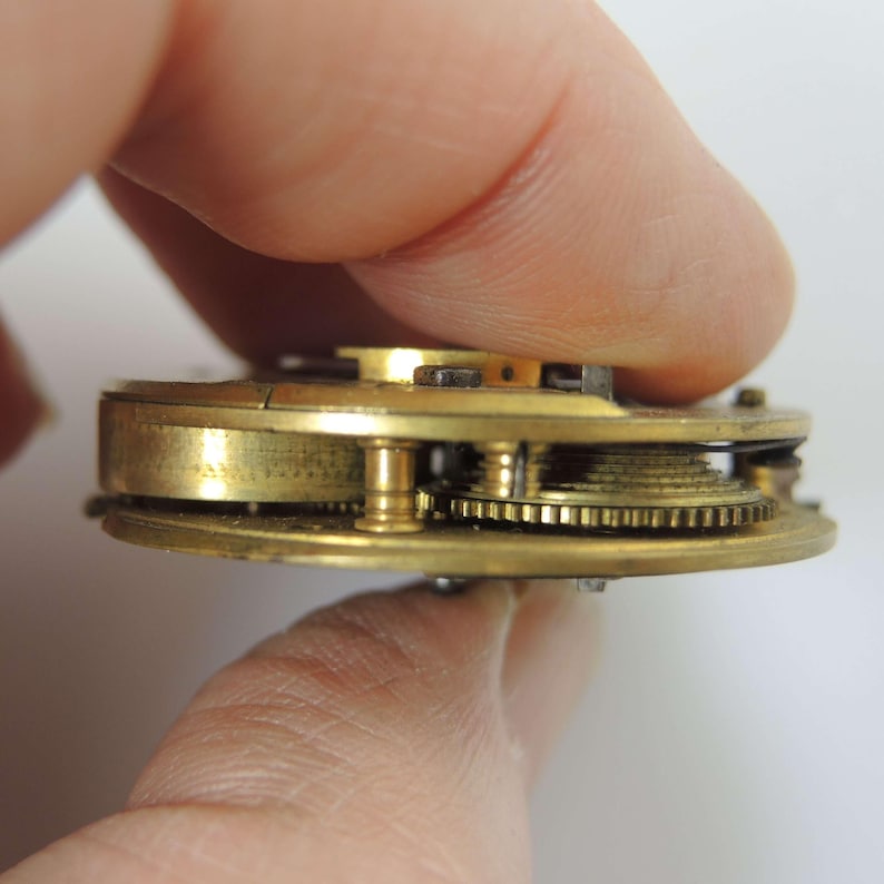 May include: Close-up of a vintage watch movement, showcasing intricate brass gears and components. The circular mechanism is held between fingers, highlighting the detailed craftsmanship and aged patina of the metal. The watch movement is a testament to precision engineering.