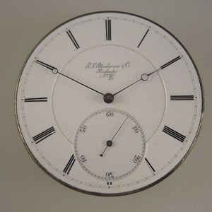 May include: A white pocket watch face with black Roman numeral hour markers and black hands. The watch face has the text "E. & J. Alexander & Co. Rochester N.Y."