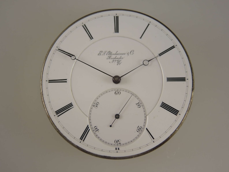 May include: A white pocket watch face with black Roman numeral hour markers and black hands. The watch face has the text "E. & J. Alexander & Co. Rochester N.Y."